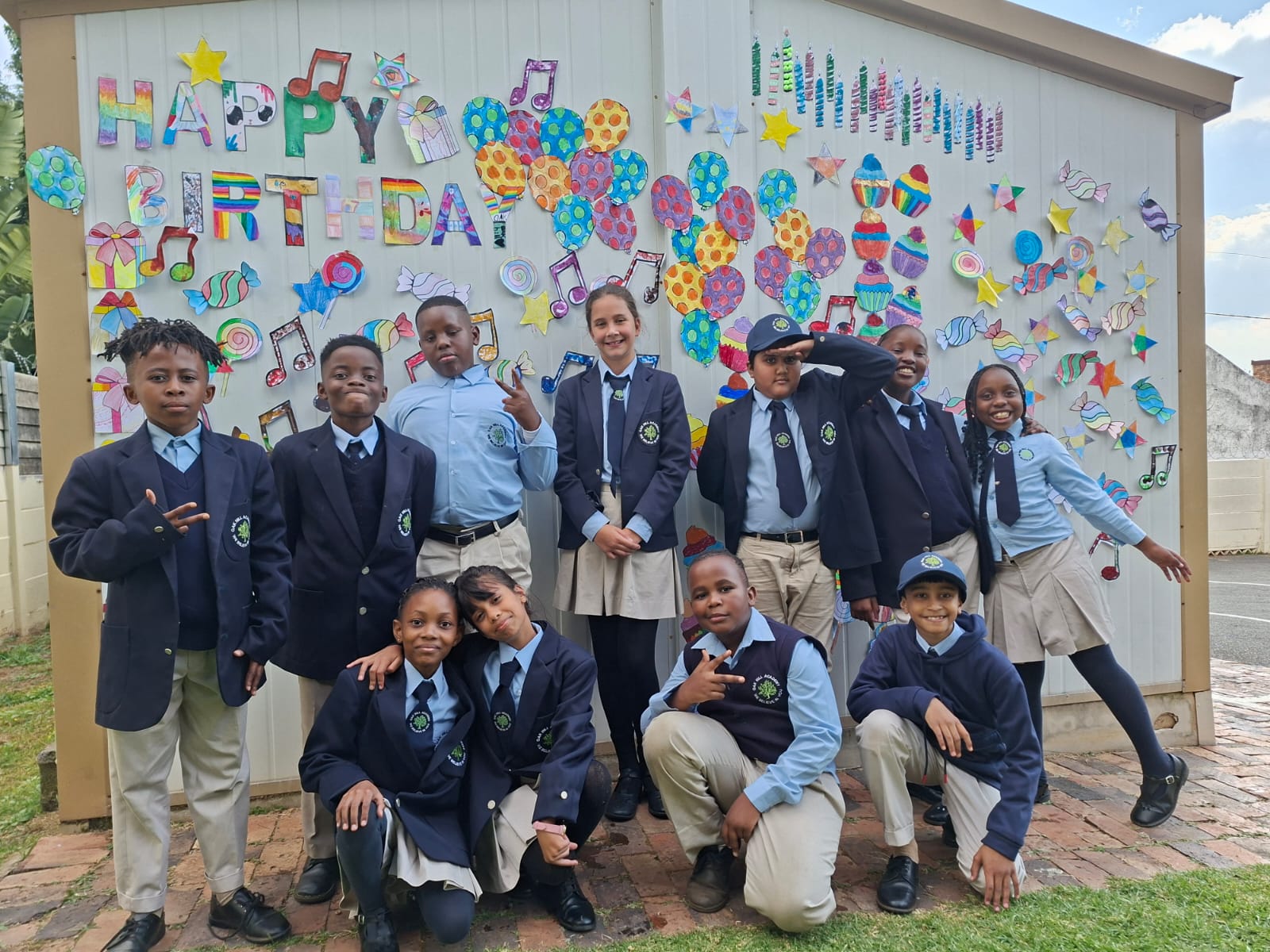 Oak Hill & Little Bean learners celebrating the schools' 25th birthday in front of a hand-decorated 'Happy Birthday' wall